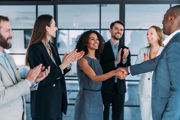 A photo in an office setting of a woman shaking hands with a man while colleagues in the background are clapping their hands in celebration.