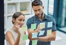 A man and a woman writing ideas on sticky notes pasted on a glass wall.