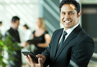 A South Asian man smiling at the camera while holding a tablet.