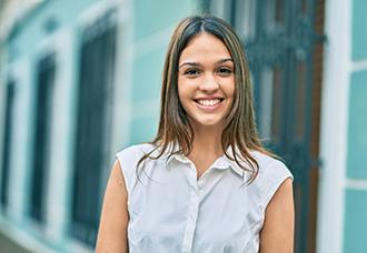 A Latino woman in a white sleeveless blouse smiling confidently while posing professionally for the camera.