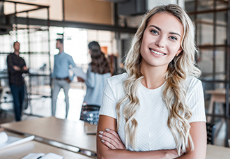 A Caucasian woman with blond hair smiling and posing for the camera, while staff members are talking with each other in the background.