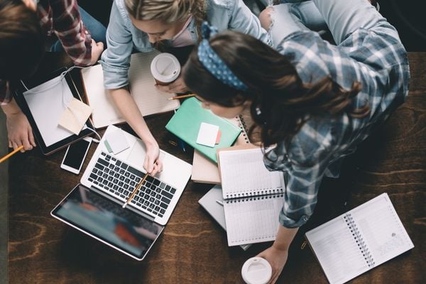 An overhead shot of students gathered around a table with notebooks, pointing with a pencil to something on a laptop, and discussing.