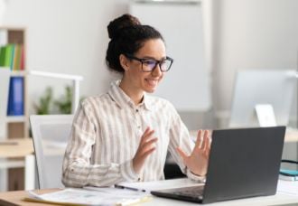 A Caucasian woman smiling while video chatting on her laptop. In the background, a bookshelf and plants are visible.
