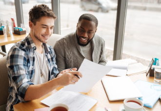 A photo of two male students smiling while discussing something. One student is helping the other student study by holding up a piece of paper with information.