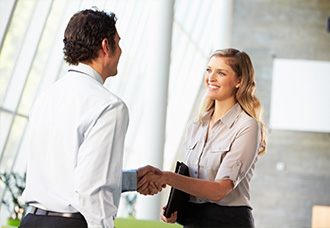 A woman and a man shaking hands in an office setting, with a professional atmosphere in the background.