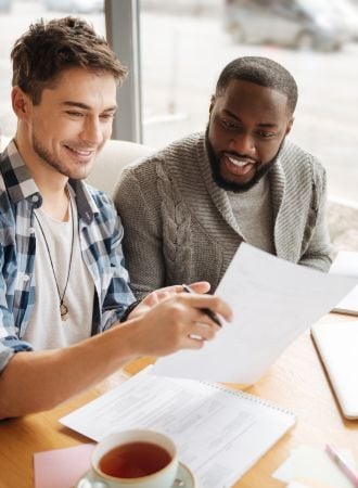 A photo of two male students smiling while discussing something. One student is helping the other student study by holding up a piece of paper with information.