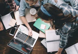 An overhead shot of students gathered around a table with notebooks, pointing with a pencil to something on a laptop, and discussing.