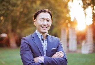 A professionally dressed Asian man with his hands folded, smiling at the camera in a natural setting.