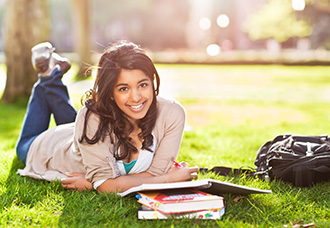 A young Asian woman is lying flat on her stomach with her hands folder and smiling at the camera. Her books and bag are on the grass near her.