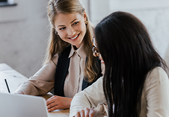 A female student smiling at another female student while both are working on something together on a laptop.