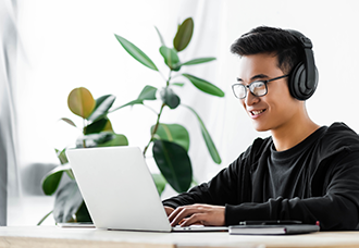 An Asian student wearing headphones and happily typing on a laptop.