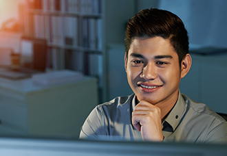 An Asian student is smiling with looking thoughtfully at his computer. The blurry background includes an open filing cabinet and a laptop placed on a desk.