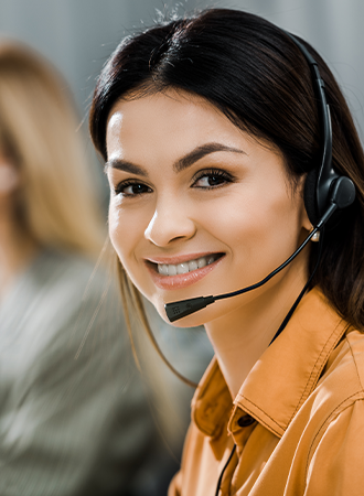 A woman with headphones smiling at the camera, with other staff members blurred in the background, indicating they are part of the contact support team.