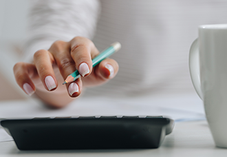 A photo capturing a close-up of a woman’s hand holding a pencil while using a calculator. A coffee mug is partially visible on the right side, adding a casual touch to the workspace setting.