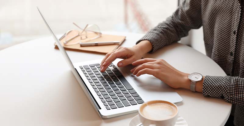 A photo of a person working on their laptop with a notebook on one side and a coffee mug on the other side.