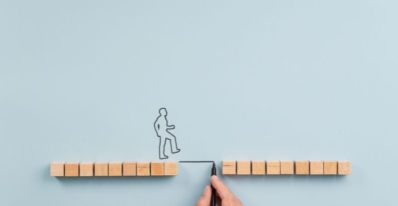 A photo featuring a blue background with wooden blocks arranged in two straight lines, leaving a gap between them. A stick figure of a person is crossing the gap using a drawn line, created by a hand holding a marker.