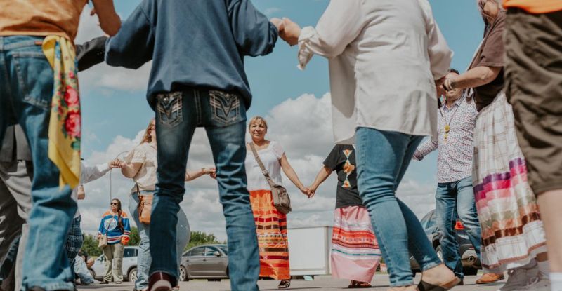 This photo was taken at an Indigenous learners gathering. Learners and staff are holding hands walking around in a circle. People are smiling with cars visible in the background. The sky looks bright and clear.