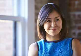 An Asian woman with short hair, wearing a blue sleeveless dress smiling confidently while posing professionally for the camera.