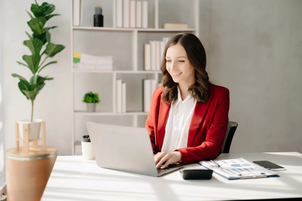 a woman in a red jacket sitting at a desk with a laptop