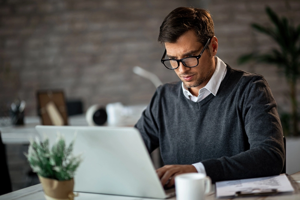 a man in a grey sweater and glasses working on a laptop