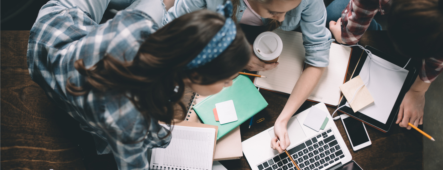 An overhead shot of students gathered around a table with notebooks, pointing with a pencil to something on a laptop, and discussing.