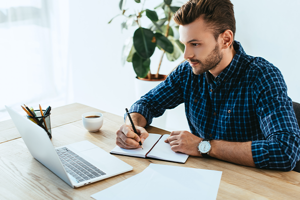 CPA Preparatory Course Learning Formats A Caucasian man wearing a blue and black checked shirt, writing in a notebook while looking at his laptop.