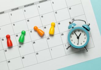 Schedules A photo of a calendar placed on a table, with magnetic pins and an alarm clock positioned on top of it.
