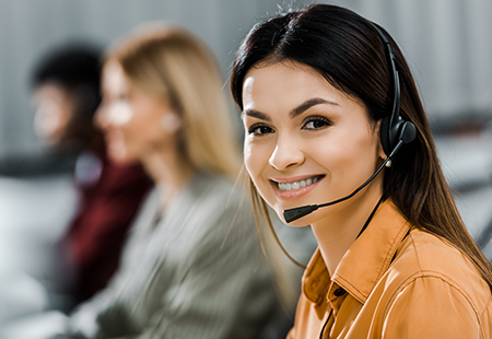 Questions? A woman with headphones smiling at the camera, with other staff members blurred in the background, indicating they are part of the contact support team.