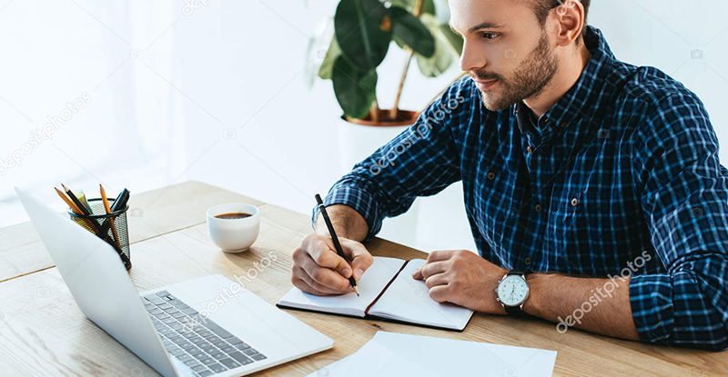 PDPA Examination A Caucasian man wearing a blue and black checked shirt, writing in a notebook while looking at his laptop.