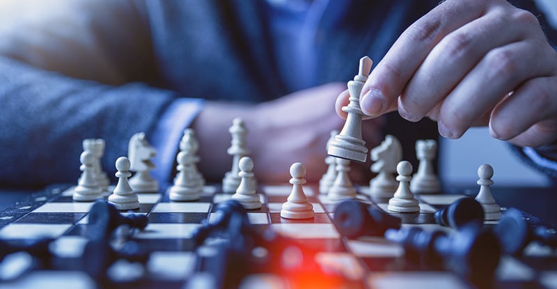 Power Move: Part 2 – The Pivot A photo of a chessboard showing a person giving a checkmate to their opponent using a white chess piece. Most of the black pieces are knocked over, while some white pieces remain standing upright.