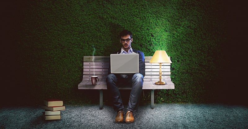 Practical tactics and time-saving tools: Part 1 A photo of a person sitting on a bench in front of a tall manicured bush with books on the ground. He can be seen typing on his laptop, a mug on his right and a lamp on his left.