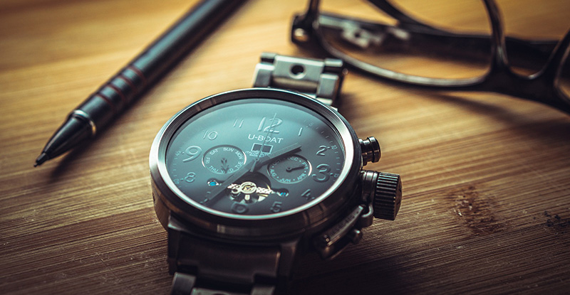 A close-up photo of a table with a watch, a pen, and glasses in focus.