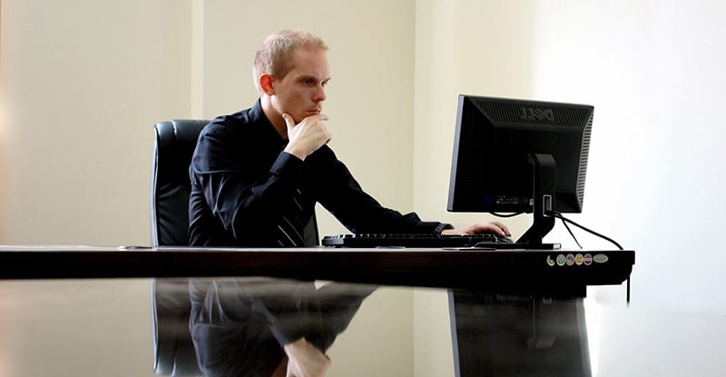 A photo of a man with his hand resting under his chin and looking at his computer. His reflection can be seen on the glass table.