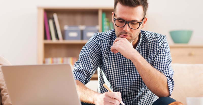 A photo of a man wearing glass has a hand under his chin while writing on a notebook with a laptop in front of him. His background features a bookshelf with filing baskets and a green bowl.