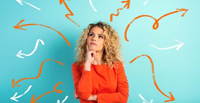 Still debating which electives to take? A photo featuring a blue background with a Caucasian woman wearing an orange shirt, looking up thoughtfully. Squiggly arrows in orange and white point away from her, symbolizing ideas or decision-making.