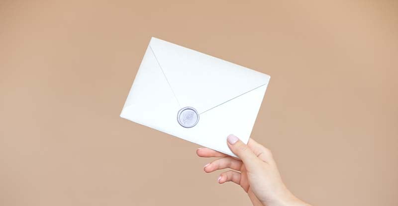 A photo featuring a light brown background with a person holding a sealed envelope.