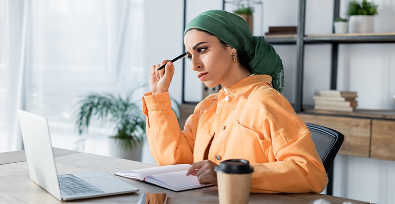 Want to become a CPA? Getting you started on your CPA path (For Canadian Accredited program applicants only) A photo of an Arab woman wearing a green turban and an orange shirt, thinking with a pencil to her head while looking at her laptop. She is seated in an office with a shelf in the background holding books and potted plants.