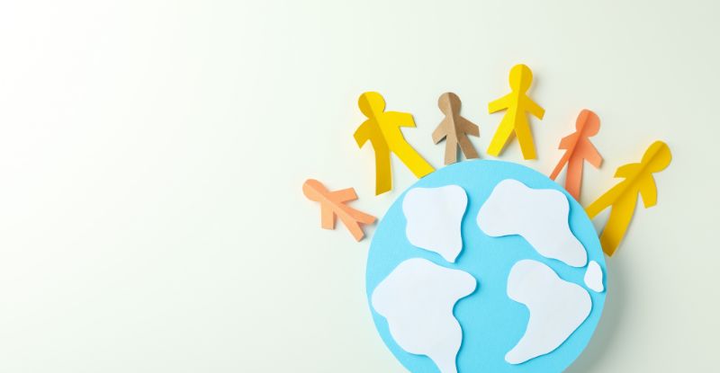 A paper cutout of a globe with small hand-holding figures of different height and colour arranged halfway around its circumference.