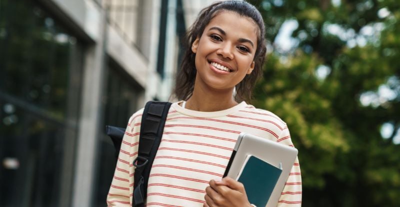 Ready to start CPA as an MOU? A photo of an African student wearing an orange horizontally striped shirt and carrying a backpack, smiling at the camera. In one hand, they hold a laptop and a notebook. The background features a university building and trees, creating an academic atmosphere.