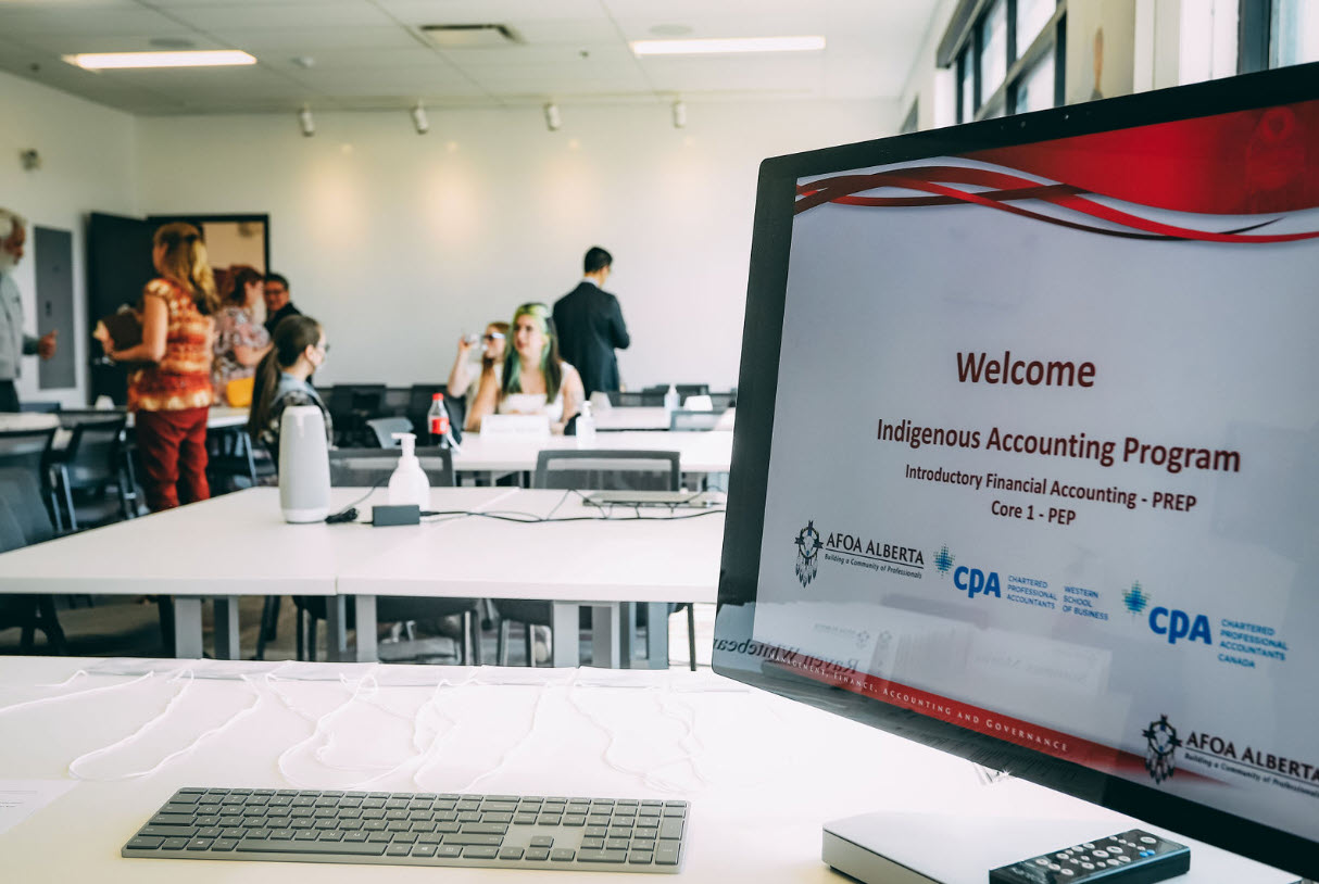 A photo from the first Indigenous Initiatives course introduction session, taken in a classroom. The camera is positioned at the teacher's desk, capturing the laptop screen and students socializing in the background.