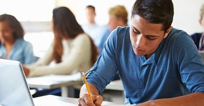What to Expect When Writing an In-Person Exam in Surpass (SecureClient) Software A photo of students seated in a classroom with the focus placed on a boy writing in a notebook with his laptop open.